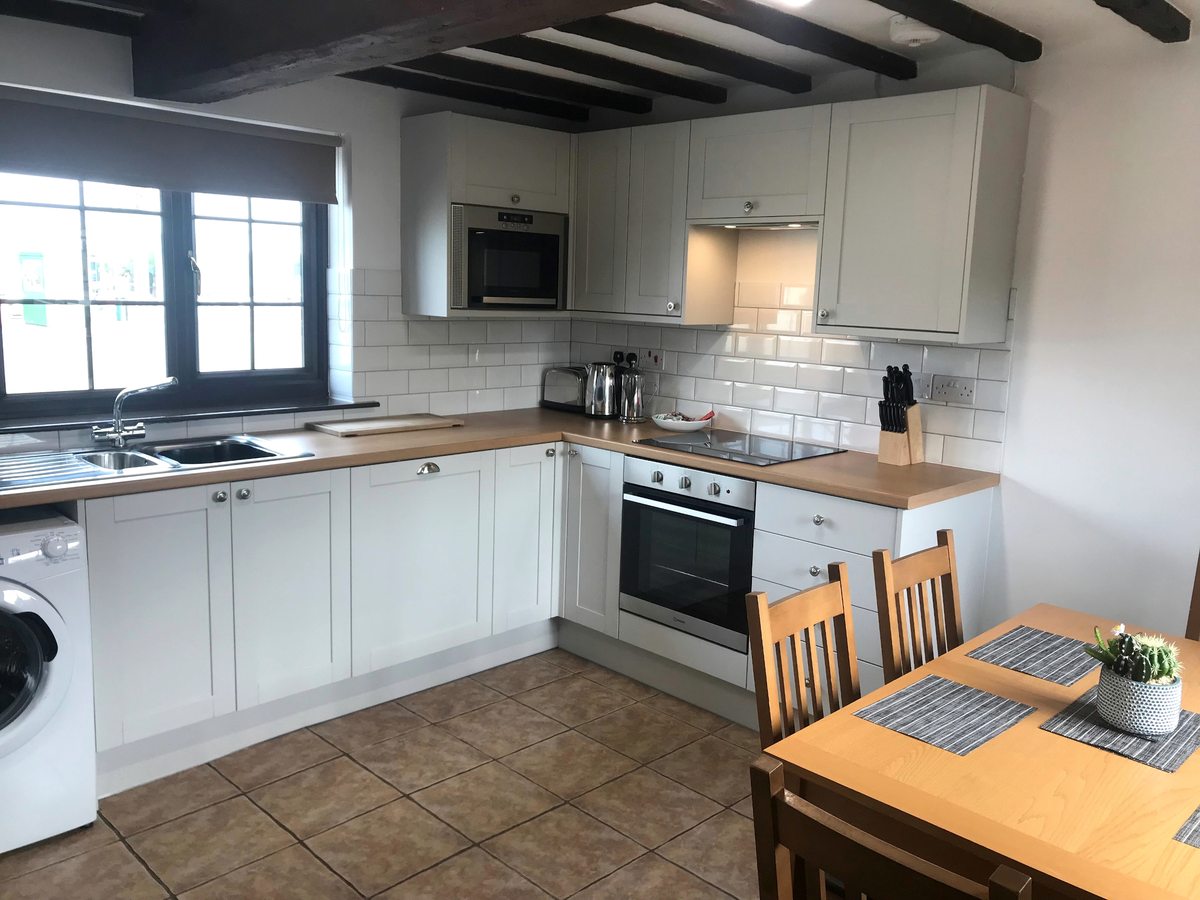 A white cottage kitchen-diner with an oven, wall units and a small wooden dining table.
