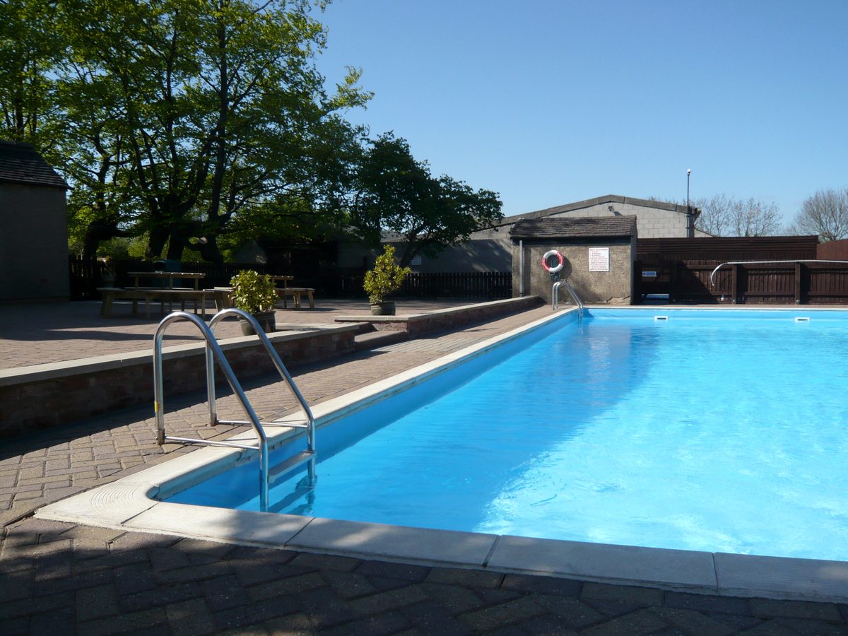 A long rectangular open-air pool with a poolside ladder and picnic benches on the paved terrace.