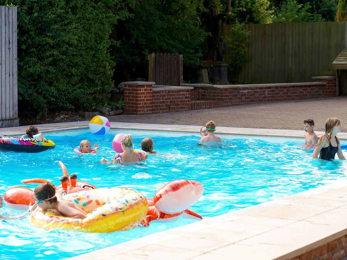 Children and adults swimming with inflatable rings and a beach ball in the open-air pool on a sunny day.