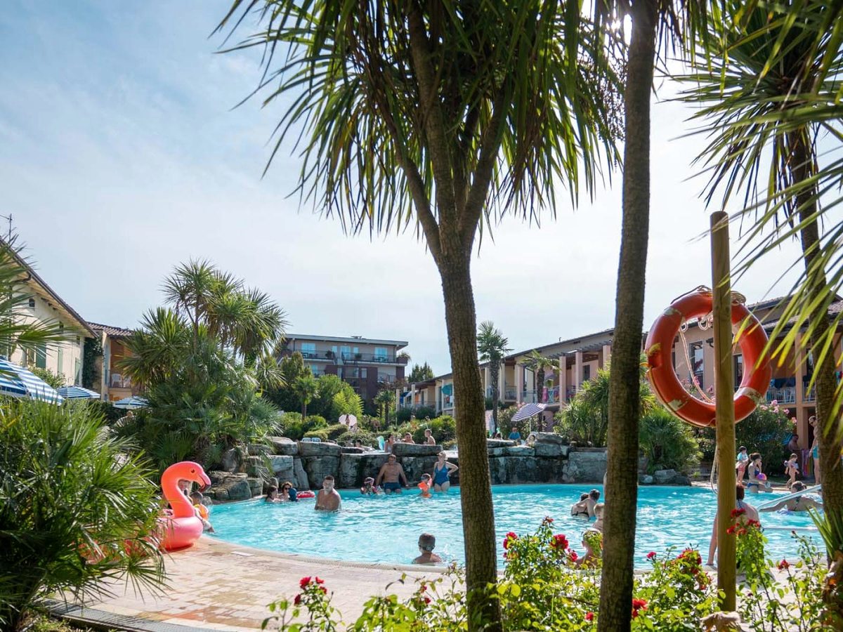 A freeform pool with rockwork and a low waterfall ringed by palms and flowering shrubs, families swimming with a flamingo float and apartment buildings behind.