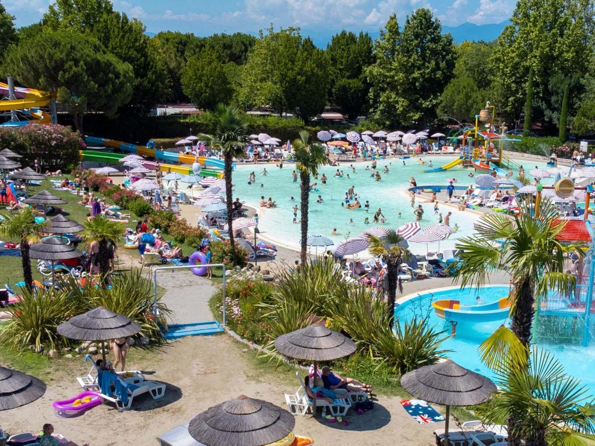An elevated view over the large freeform main pool with a beach-style entry, thatched parasols and sun loungers, with waterslides to one side and a splash tower to the other.
