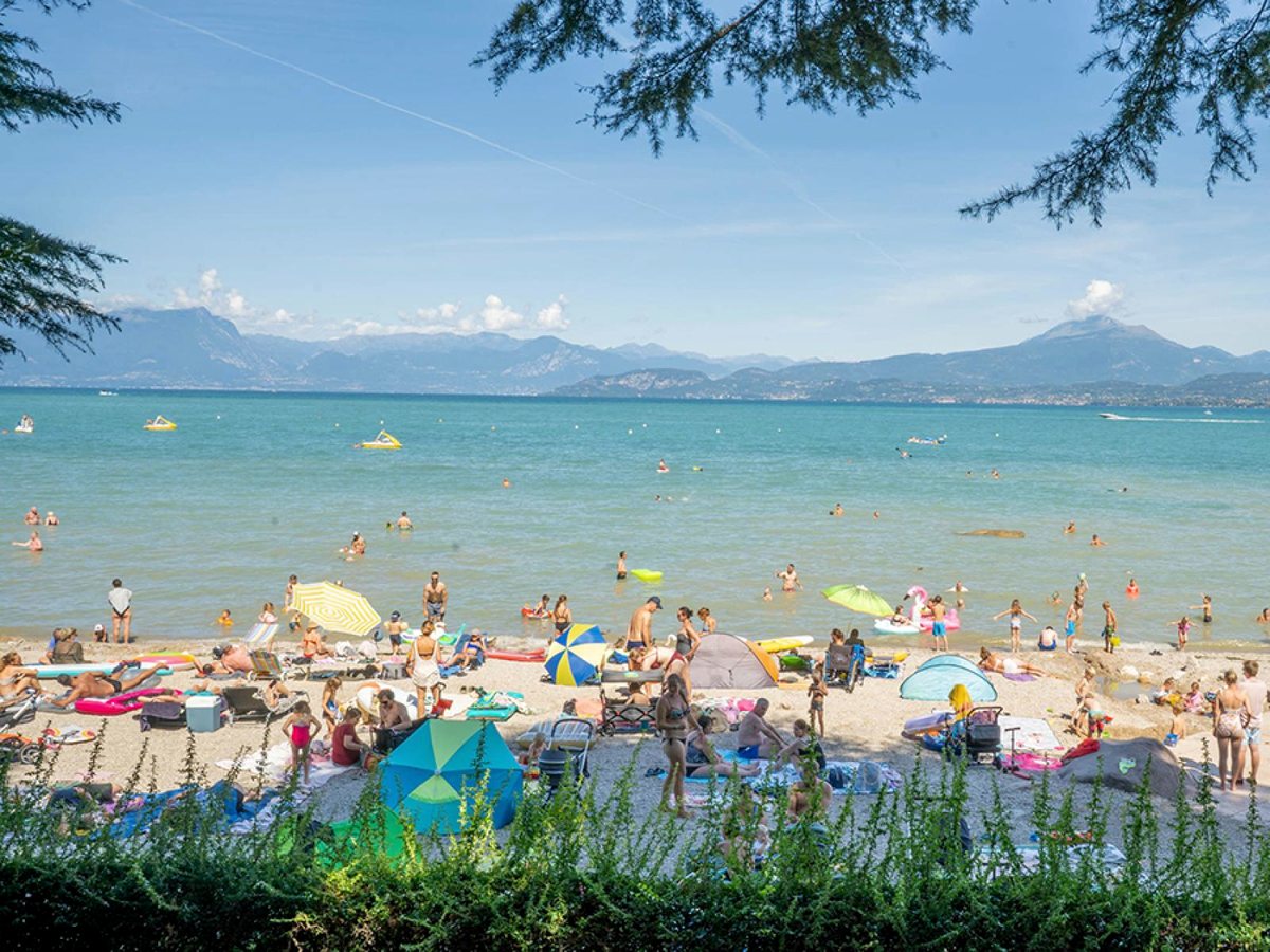 A busy shingle lakeside beach crowded with parasols, inflatables and swimmers, with wooded hills across the water.