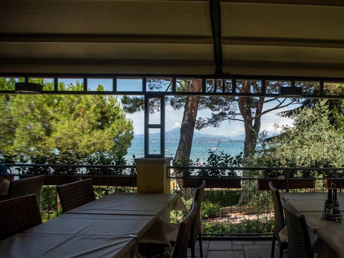A covered restaurant terrace with laid tables looking out through pines to Lake Garda and the mountains beyond.