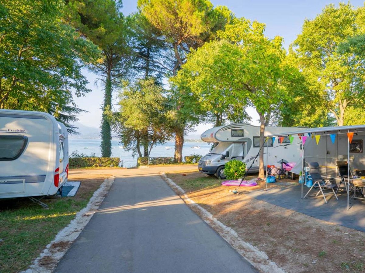 Tree-shaded touring pitches with a caravan and a motorhome, camping chairs and bunting, with Lake Garda visible through the trees.