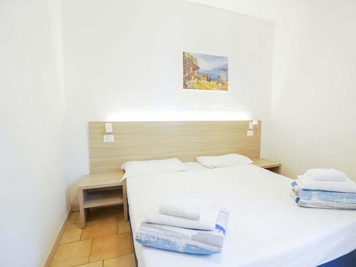 A compact double bedroom with a pale wood headboard, bedside reading lights, folded towels and a framed landscape print.