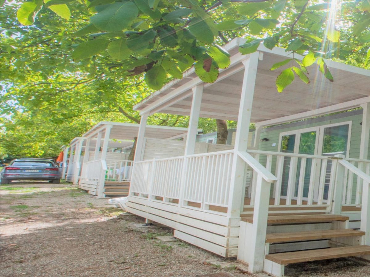 A row of white mobile homes with covered verandas and steps, shaded by mature trees, with a car parked on the access lane.