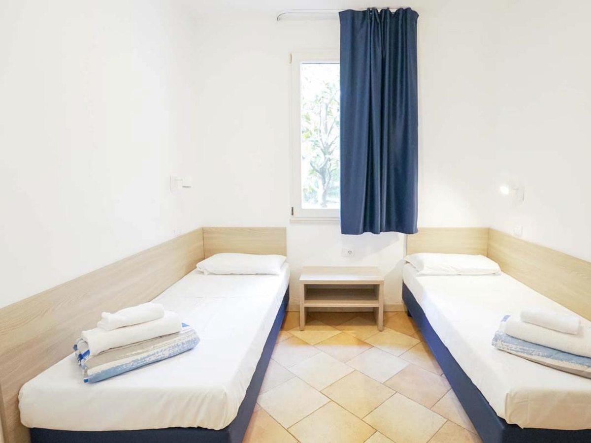 A twin bedroom with two single beds either side of a shared bedside table, a window with a blue curtain and a tiled floor.