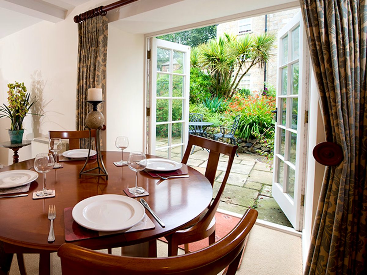 A cottage dining room with a round wooden table laid for six and open french doors onto a paved garden terrace.