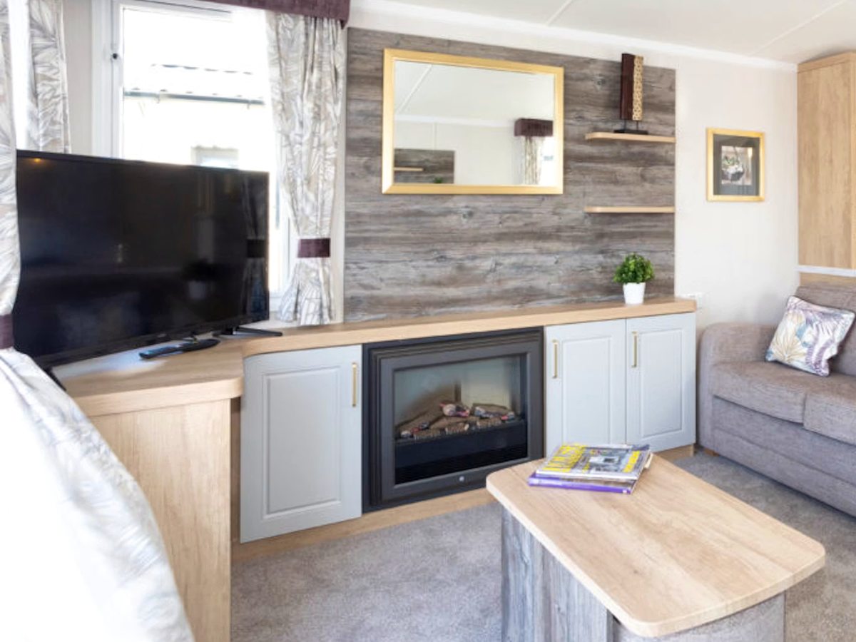 A static caravan lounge with a grey corner sofa, a wall-mounted flat-screen TV, an electric fireplace and built-in storage.
