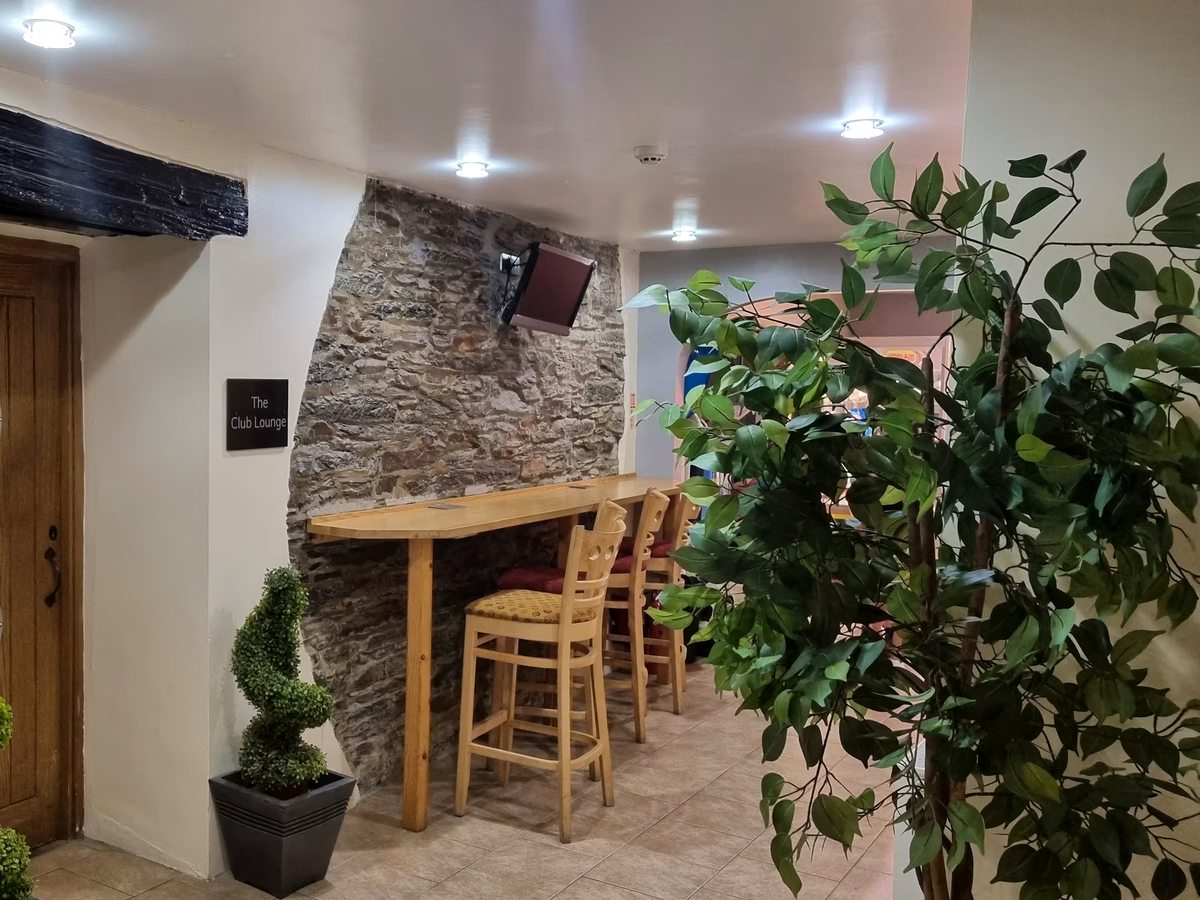 The Club Lounge, a seating nook with a bar-height wooden counter and stools against an exposed stone wall, a tiled floor and a large potted plant.