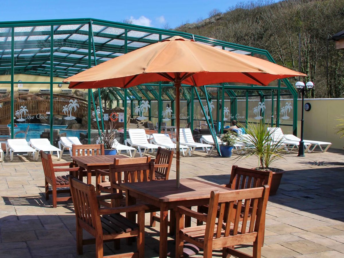 An outdoor pool patio with wooden dining sets, orange parasols and white sun loungers on paving beside the green-framed pool building, a wooded hillside behind.
