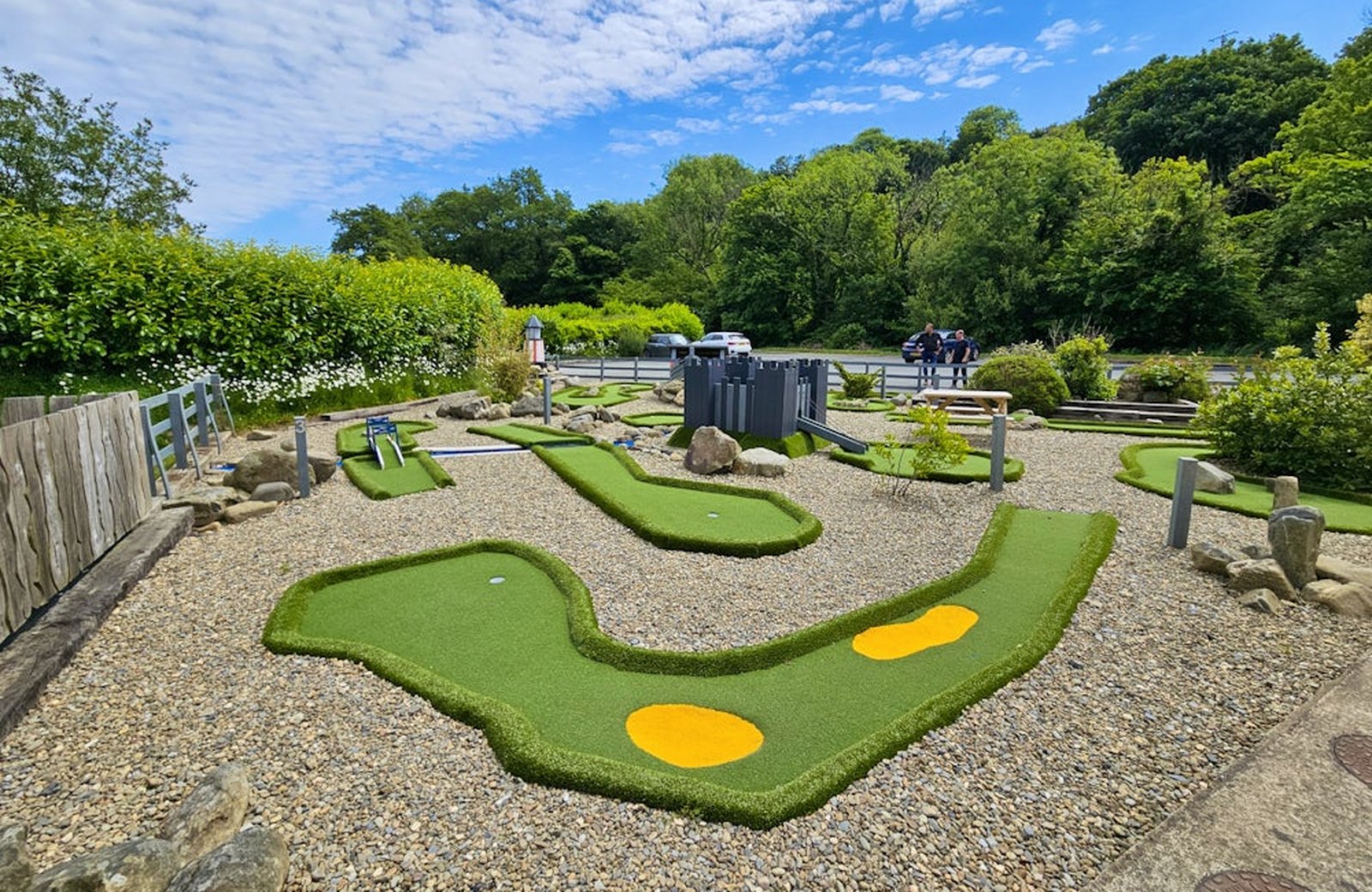 The crazy golf course at Cardigan Bay Holiday Park, its bright green putting lanes set on gravel with a wooded hillside behind under a blue sky.