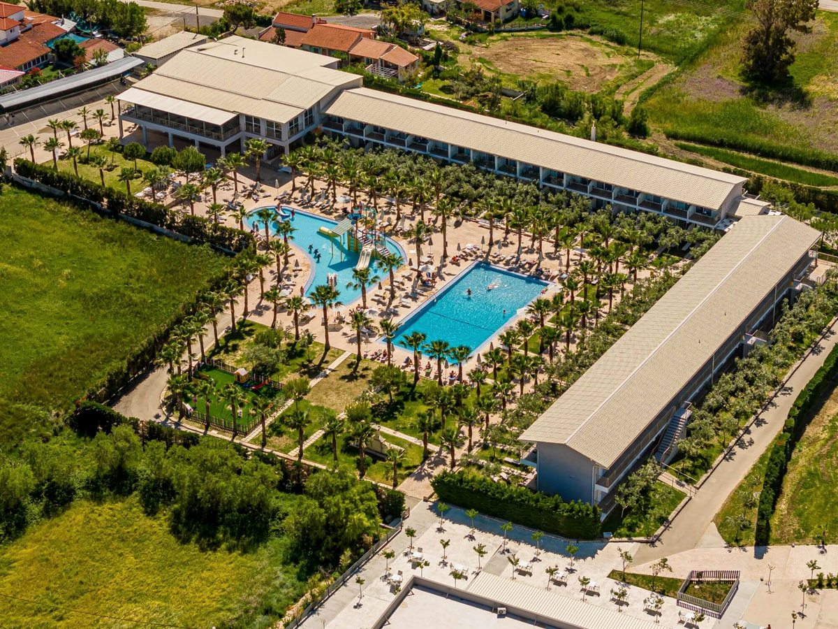 Aerial view of a rectangular pool and a smaller children's pool among palm trees, enclosed by two-storey white resort buildings.