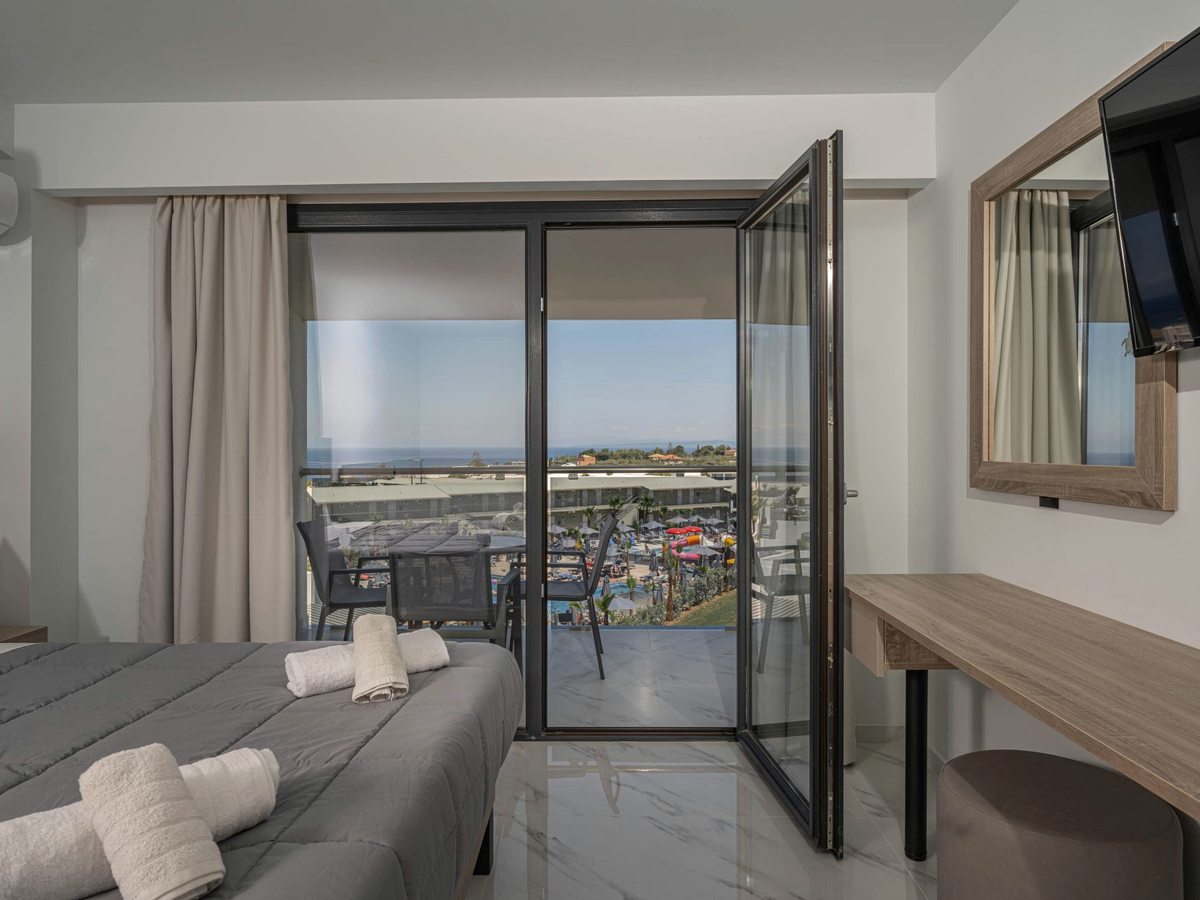 Hotel room with a double bed, a desk and a wall-mounted TV, the glass balcony door open to a view over the waterpark and the sea beyond.