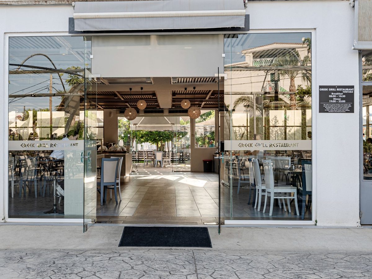 Glass-fronted entrance to the Greek grill restaurant, with tables visible inside beyond a level threshold.