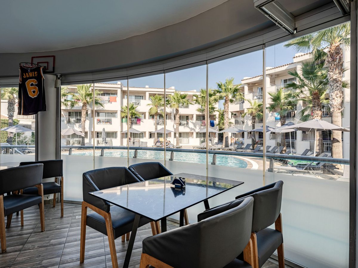 Indoor bar lounge with black leather chairs at a marble table beside curved floor-to-ceiling windows overlooking the pool.