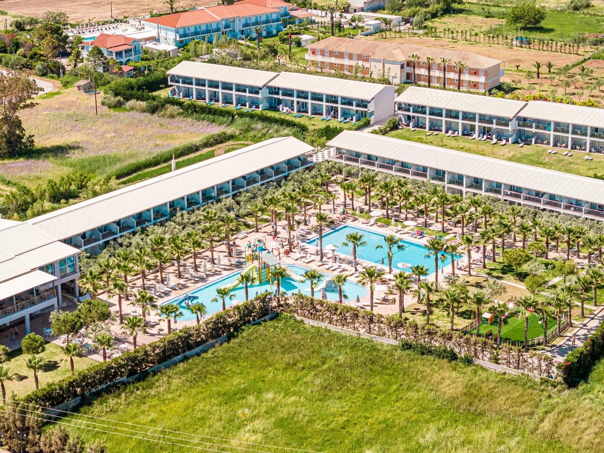 Aerial view of long two-storey white resort buildings around a palm-filled pool area, with farmland beyond.