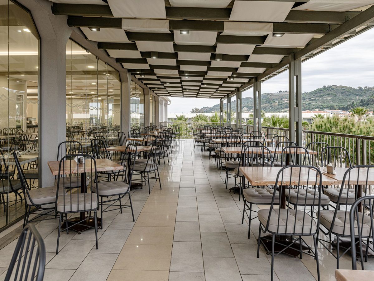Long covered dining terrace with metal chairs at tables along a tiled floor, open to a view at the far end.