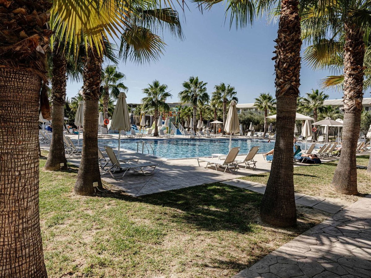 Rectangular pool with sun loungers and parasols seen through tall palm trees on a lawn, white resort buildings behind.