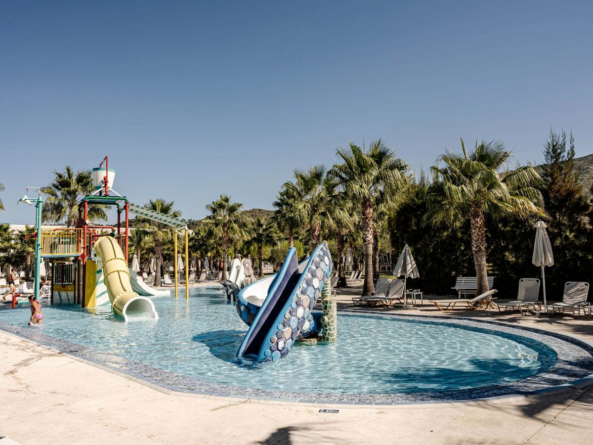 Shallow children's pool with a play structure and small tube and mat slides, palm trees and a hillside behind.
