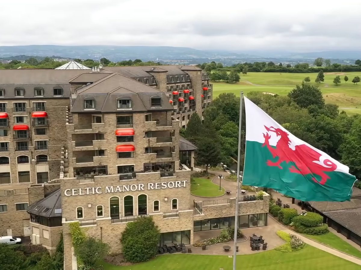 An aerial view of the stone-built Celtic Manor Resort Hotel beside a golf course, with a large Welsh flag flying and the Usk Valley behind.