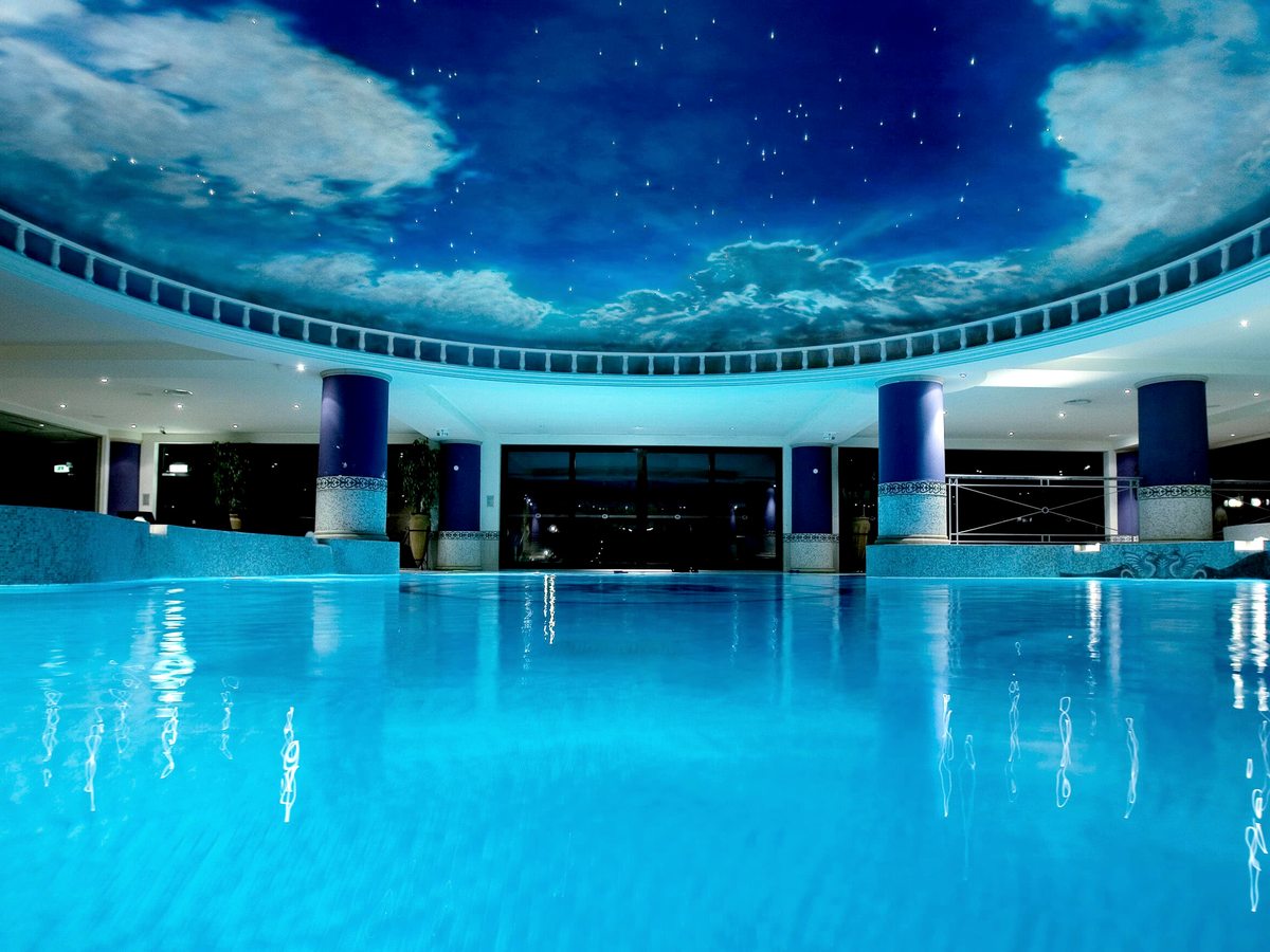 A large indoor pool with blue water beneath a domed ceiling painted as a night sky, ringed by columns and a curved upper balcony.