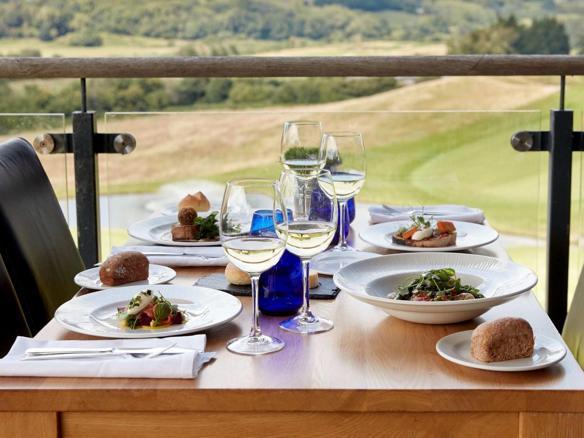 A table laid for lunch on a railed terrace with wine and water glasses, looking out over the golf course and countryside.