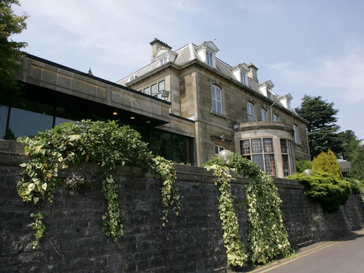 The ivy-clad stone facade of the 19th-century Manor House, with dormer and bay windows above a stone retaining wall.