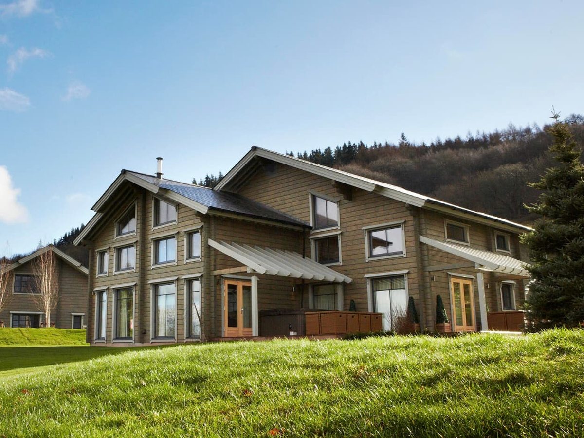 A timber-clad Hunter Lodge with large windows and a pitched roof on a grassy hillside backed by woodland.