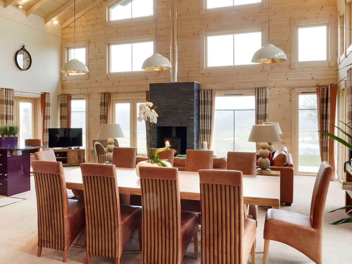A double-height timber lodge interior with a long dining table and chairs, sofa seating and a fireplace beneath large windows.