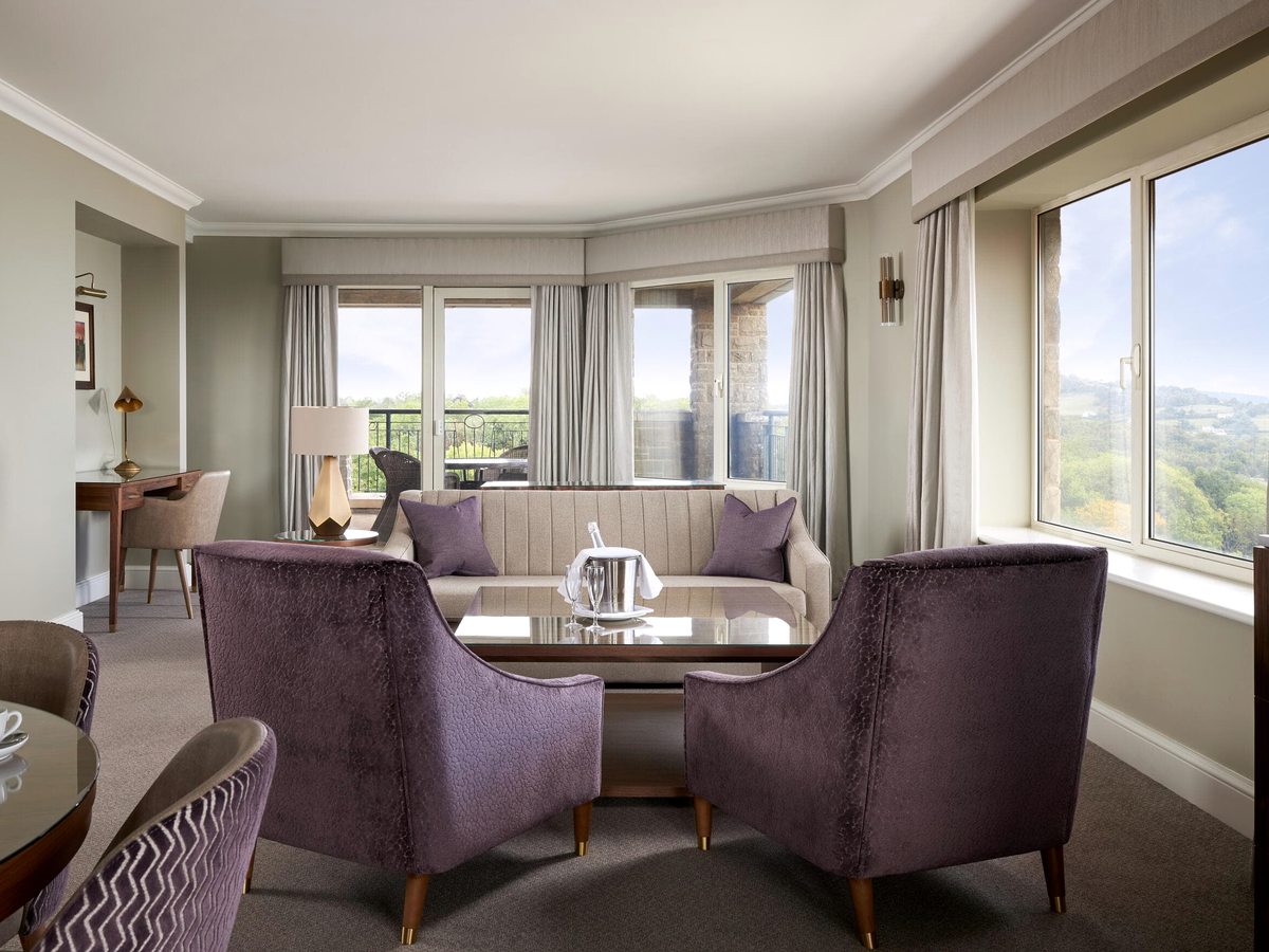 A suite sitting room with upholstered armchairs, a coffee table and a writing desk beneath panoramic windows onto open countryside.