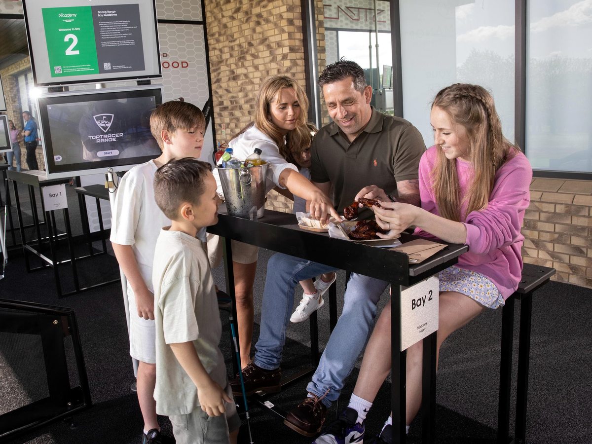 A family gathered around a table at a covered Toptracer golf bay with a touchscreen at the Golf Academy, eating together.