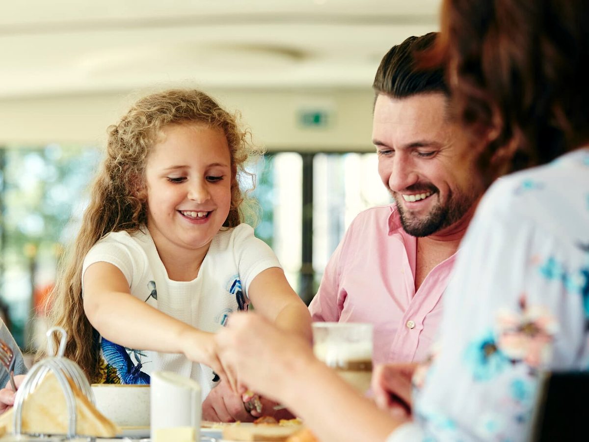 A father and young daughter smiling across a restaurant table set with plates and glasses in a daylit dining room.