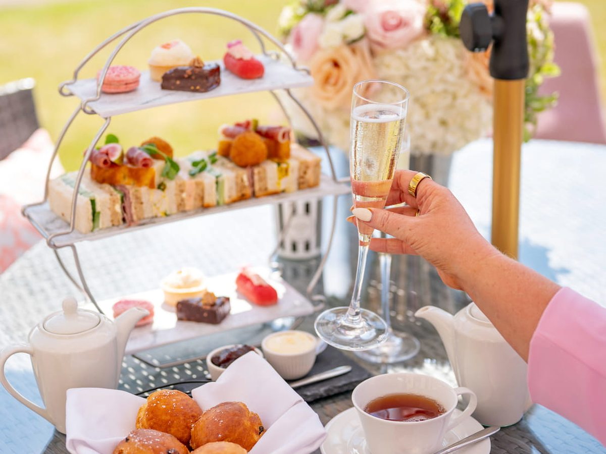 A three-tier stand of finger sandwiches, scones and cakes on a terrace table with a teapot, teacups and a parasol.
