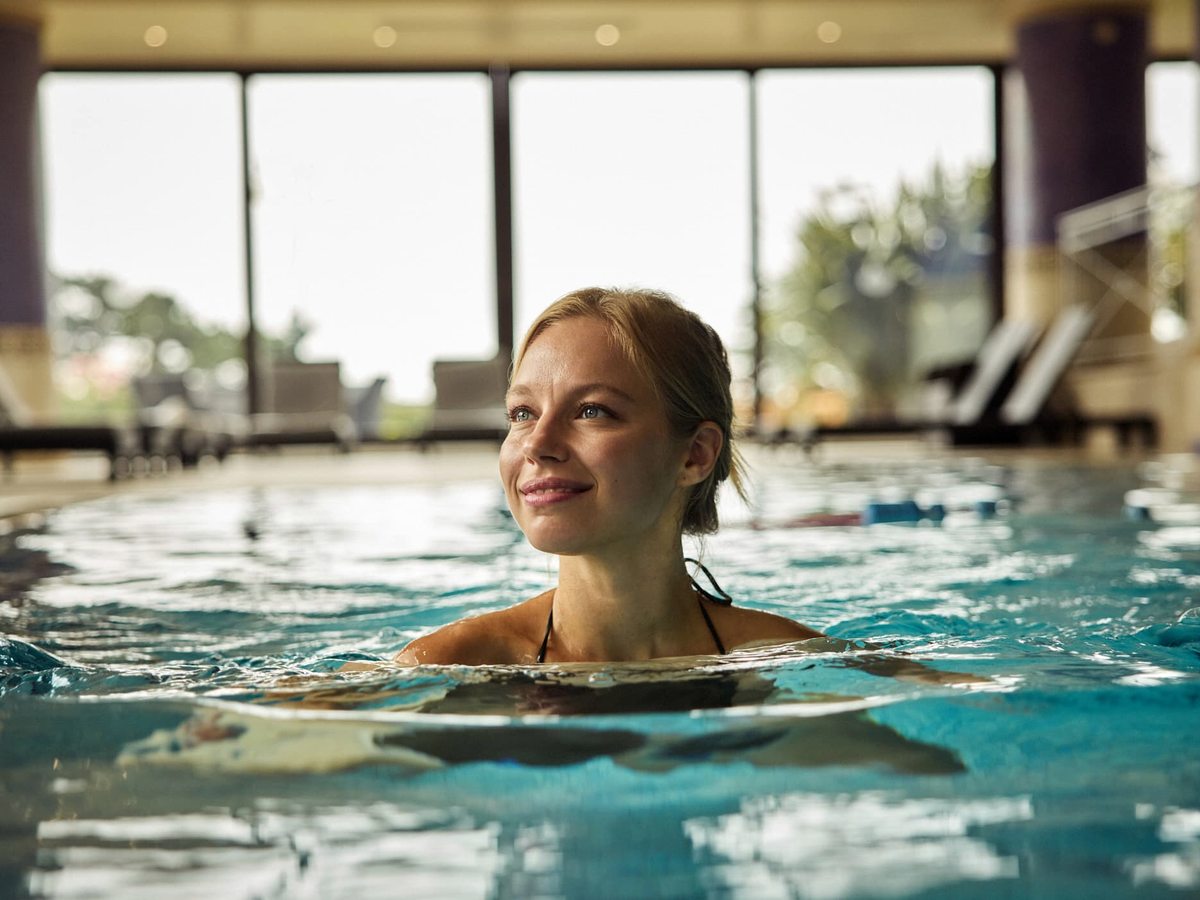 A swimmer in a bright indoor pool with poolside loungers and large windows letting in daylight.