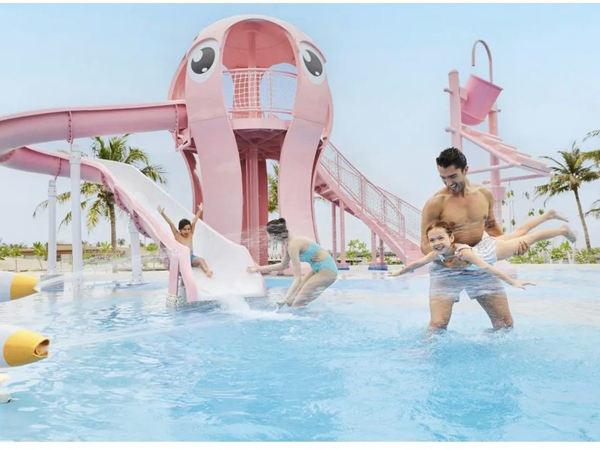 A pink octopus-shaped water tower with slides above a shallow splash pool, children and a parent playing, palm trees behind.