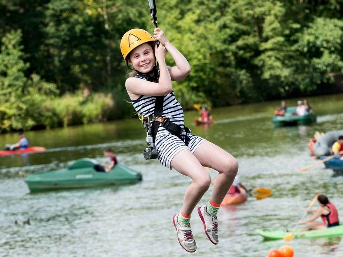 A child in a yellow helmet riding a zip wire above the lake, with pedalos and swimmers on the water below.