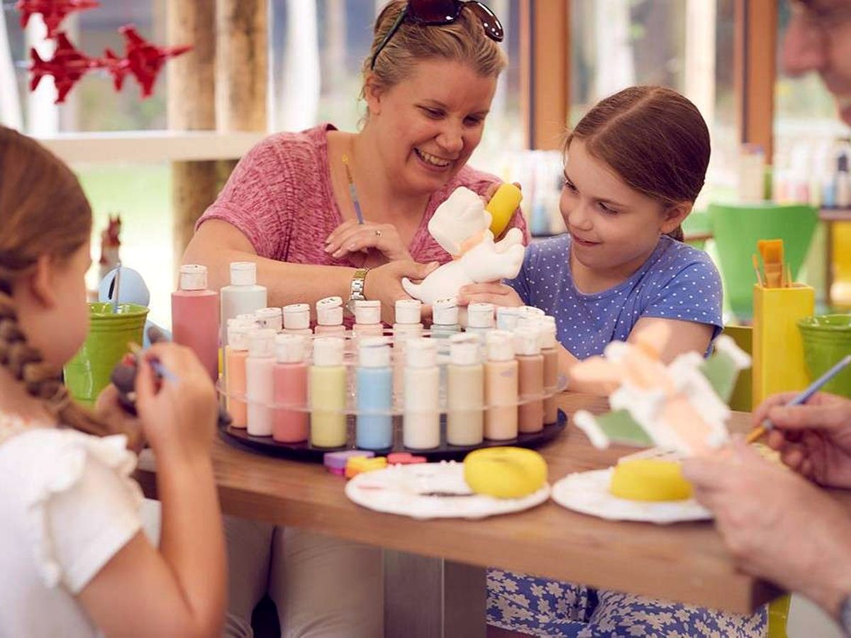An adult and two children painting ceramic pieces at a table lined with pots of coloured paint in a bright craft room.