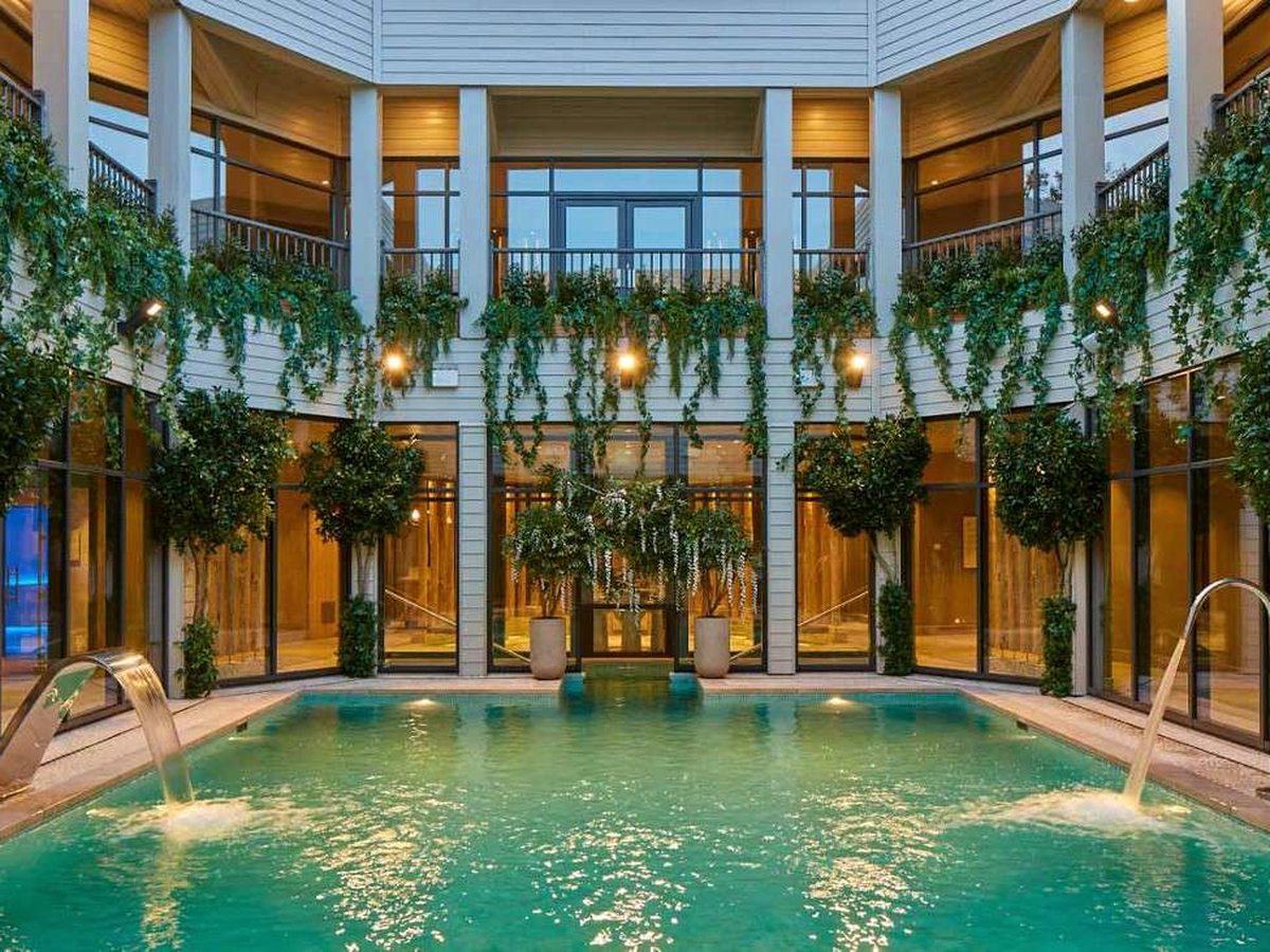 A rectangular green-lit pool in a timber courtyard, with two arching water spouts, clipped bay trees and trailing greenery along the balconies.