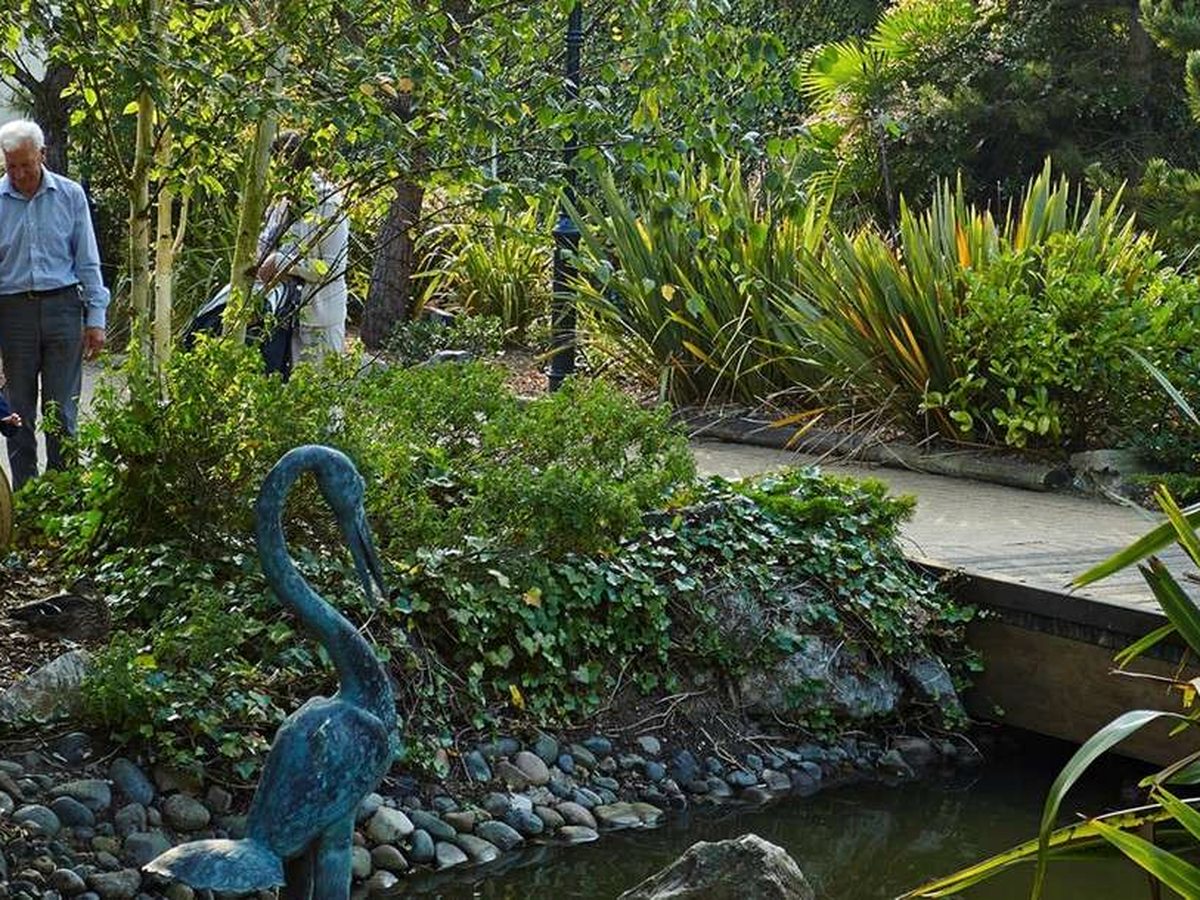 A planted garden of grasses, palms and shrubs around a pebble-edged pond, with a heron sculpture and a paved path where two people are walking.