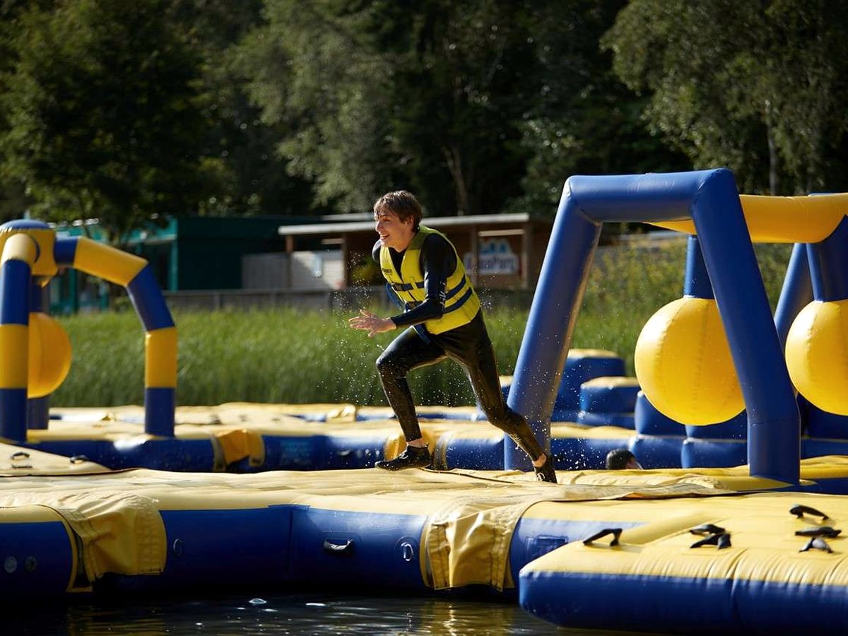 A man in a wetsuit and buoyancy aid running across a blue and yellow inflatable obstacle course floating on the lake.