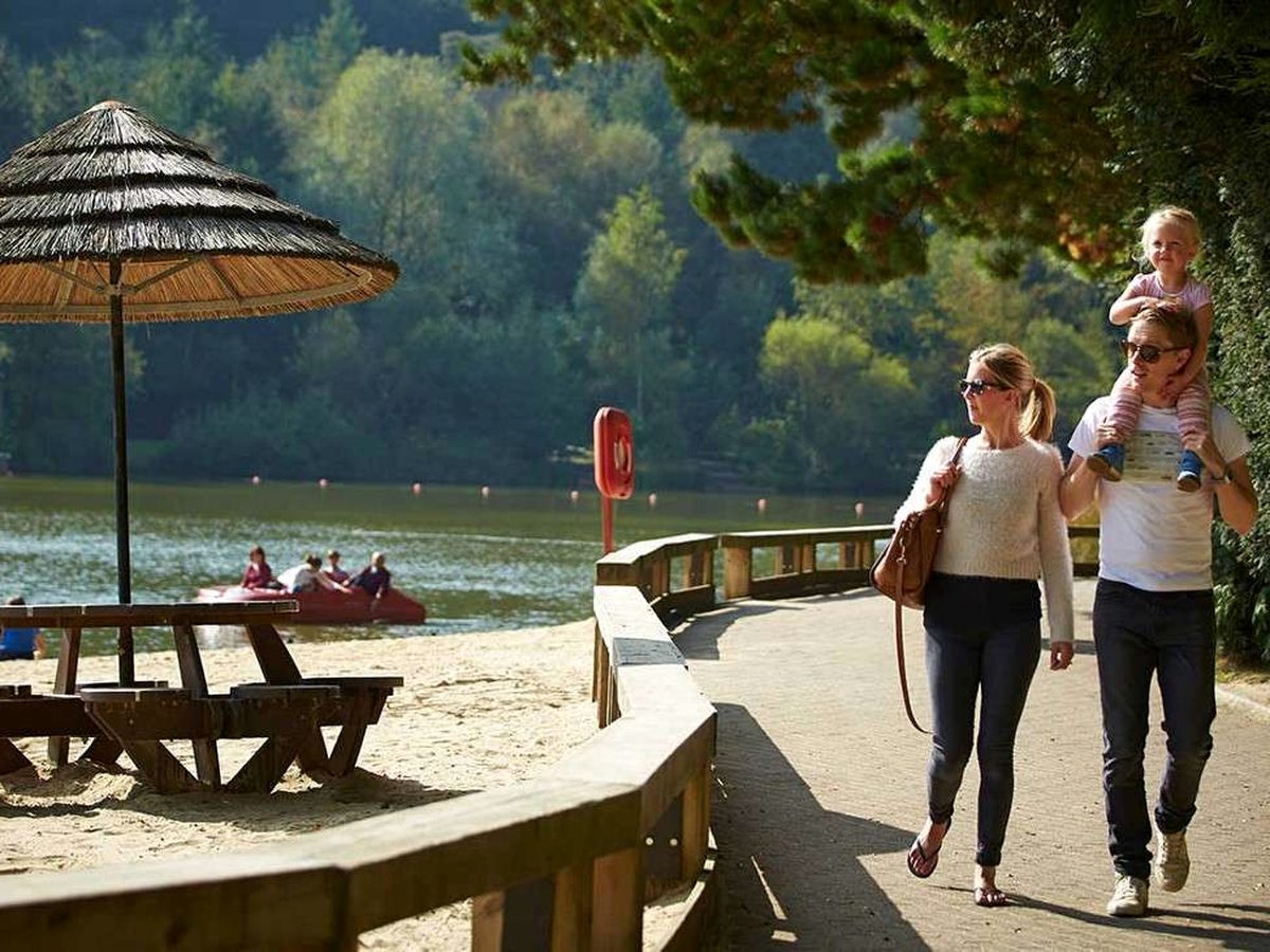 A family walking along a paved path beside a sandy lakeside beach with picnic tables and a thatched parasol.