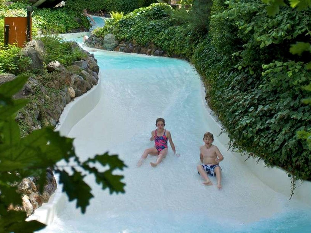 Two children sitting in the shallow rushing water of an outdoor rapids channel banked by rocks and greenery.