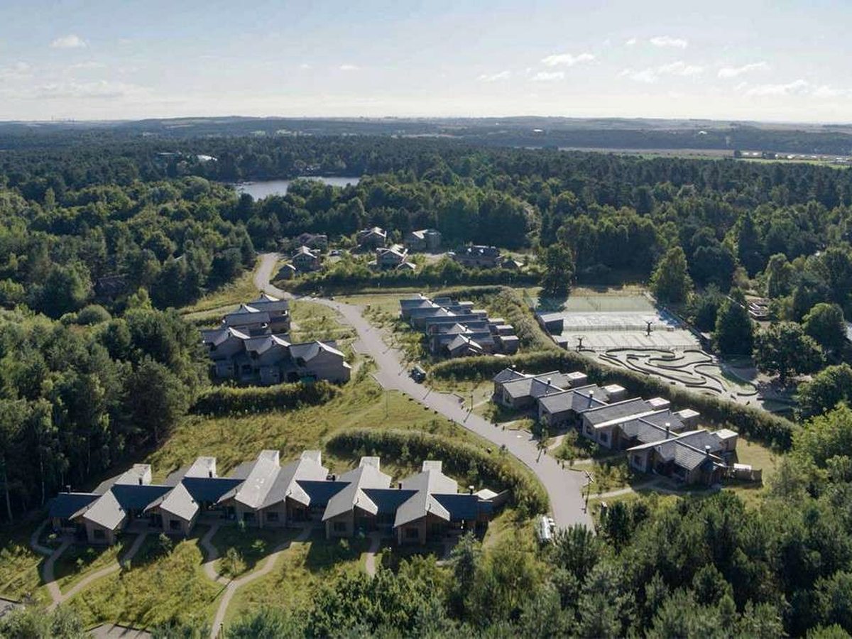 Aerial view of rows of pitched-roof holiday lodges set among dense green woodland, with curving paths, a tennis court and a lake in the distance.