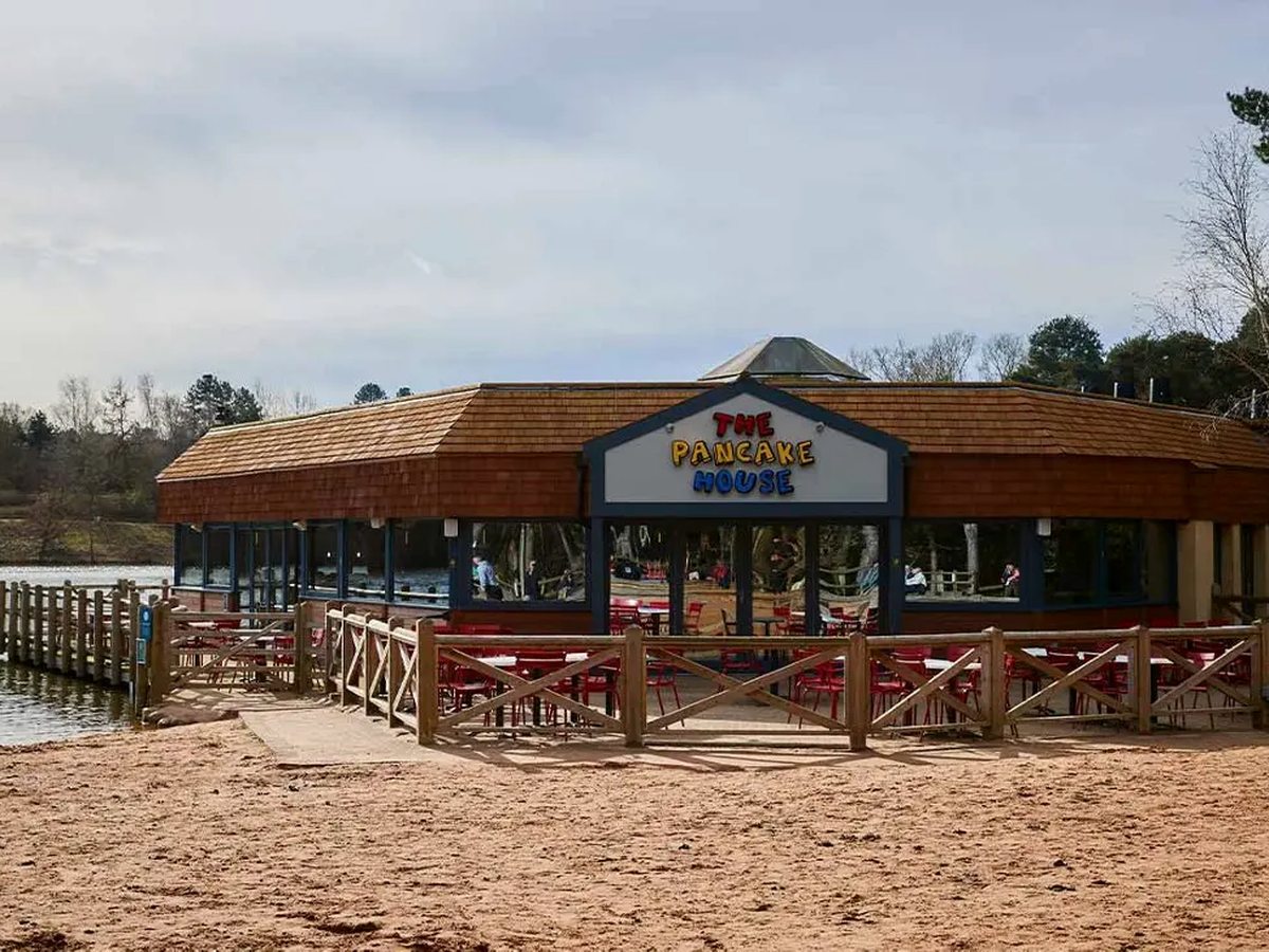 A single-storey timber restaurant with a shingled roof and a bright Pancake House sign, fronted by red outdoor chairs behind a wooden fence on a sandy lake shore.