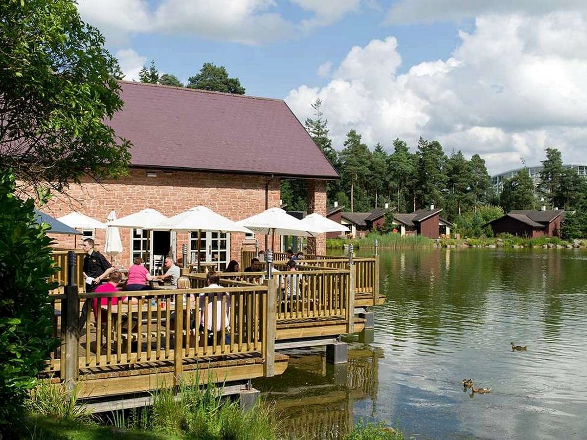 A red brick pub with a wooden deck built out over the lake, families eating under white parasols and lodges among the pines on the far bank.