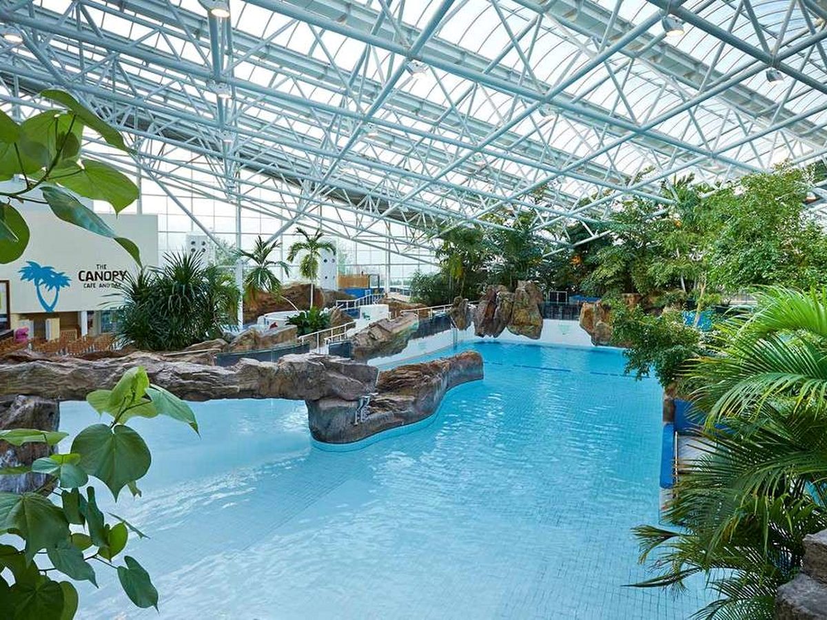 A pale blue indoor pool edged with artificial rocks and tropical planting under a high glazed roof, with a poolside cafe and bar sign on the left.