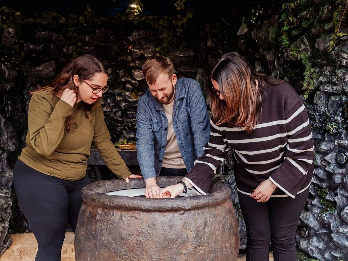 Three adults leaning over a large stone urn working out a puzzle in a themed room with rough stone walls and trailing ivy.