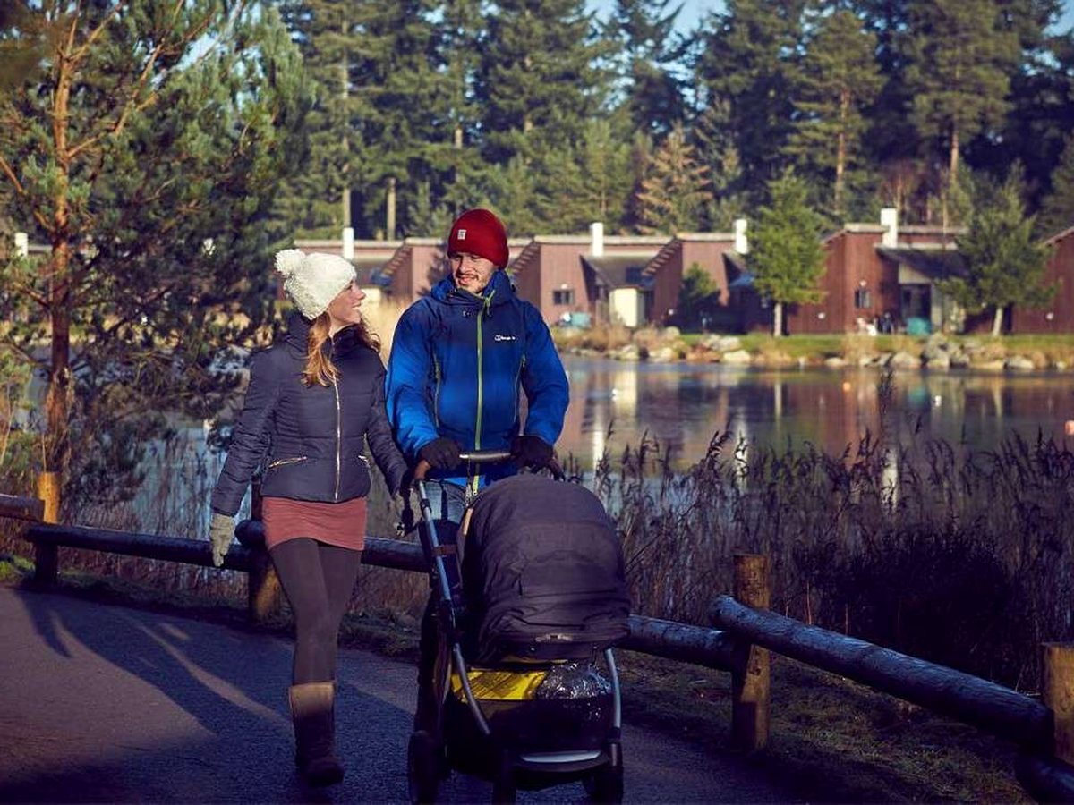 A couple in winter coats pushing a pram along a paved path beside the lake, with a wooden rail, reeds and timber lodges in the pine forest across the water.