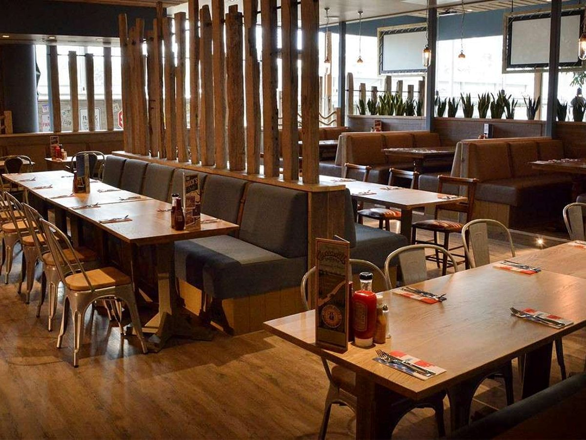 A restaurant dining room with long wooden tables, grey banquette benches, metal chairs and timber post screens, laid up with cutlery and condiments.